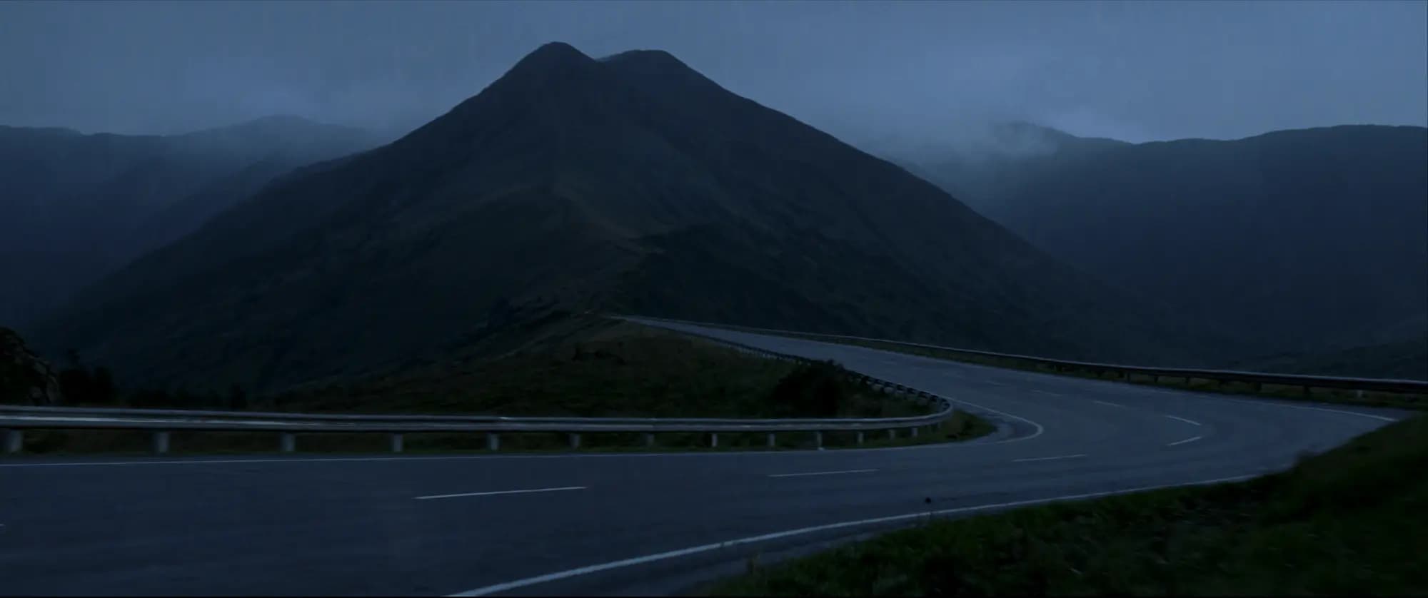 Carretera de montaña al anochecer frente a un pico cónico, con niebla baja. Sin vehículos ni personas.